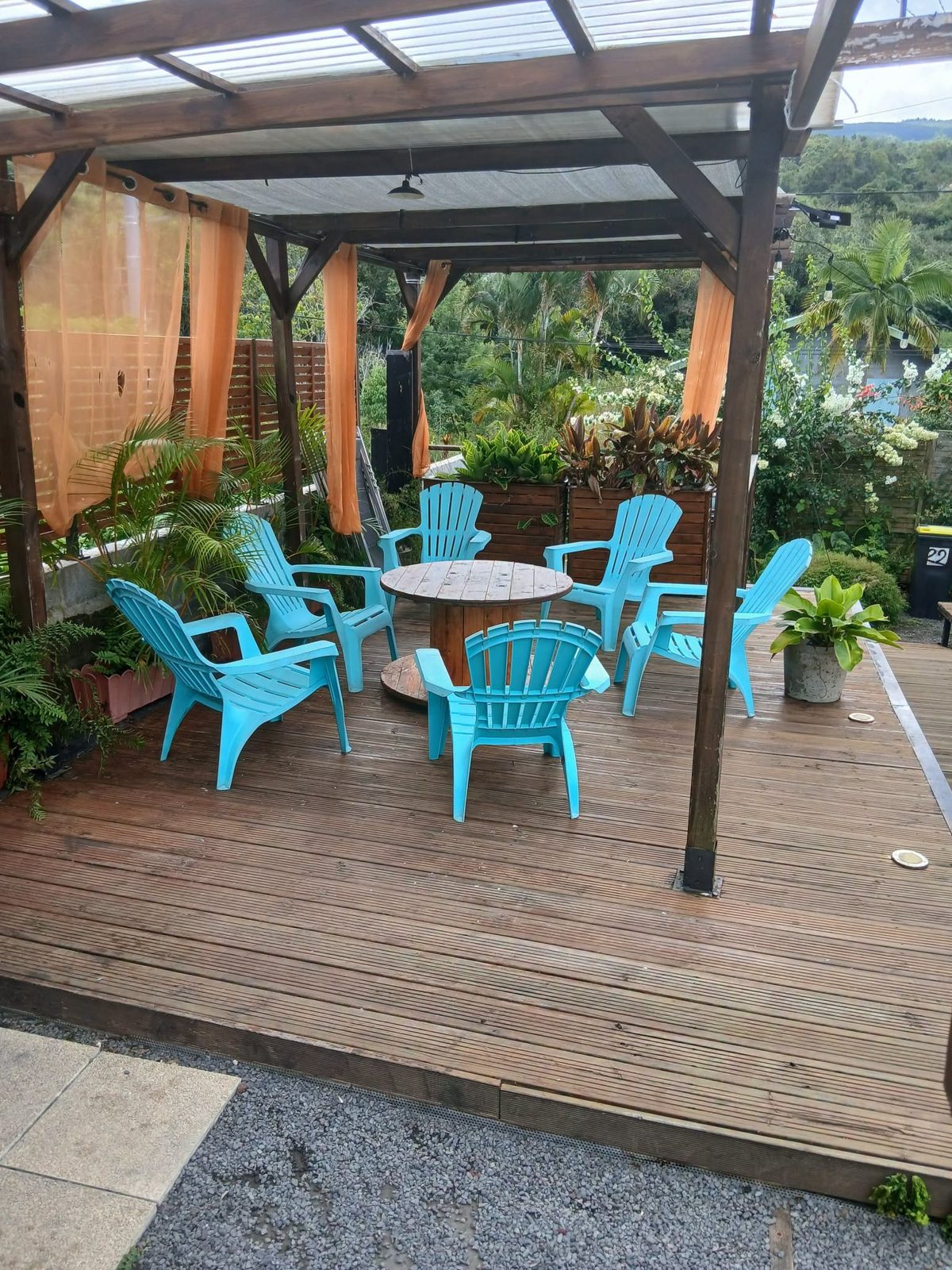 Terrasse en bois couverte avec chaises turquoise, rideaux orange et jardin tropical